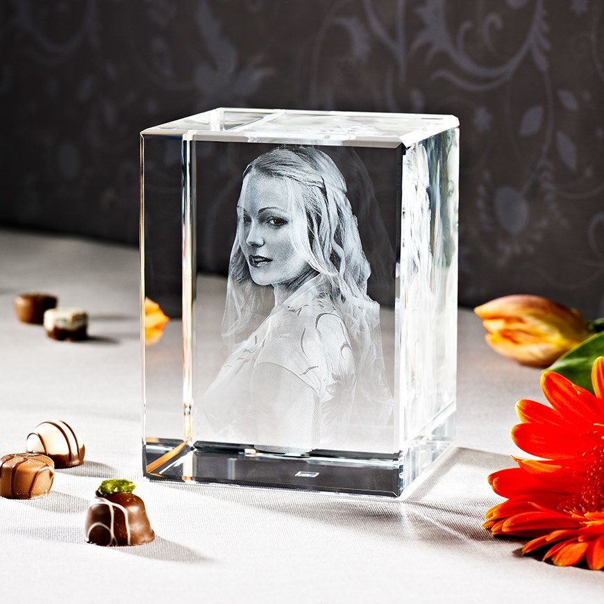 Kristallwürfel mit 3D-Lasergravur einer Frau mit langen Haaren und einem Kleid, umgeben von Pralinen und orangefarbenen Blumen auf weißem Tuch