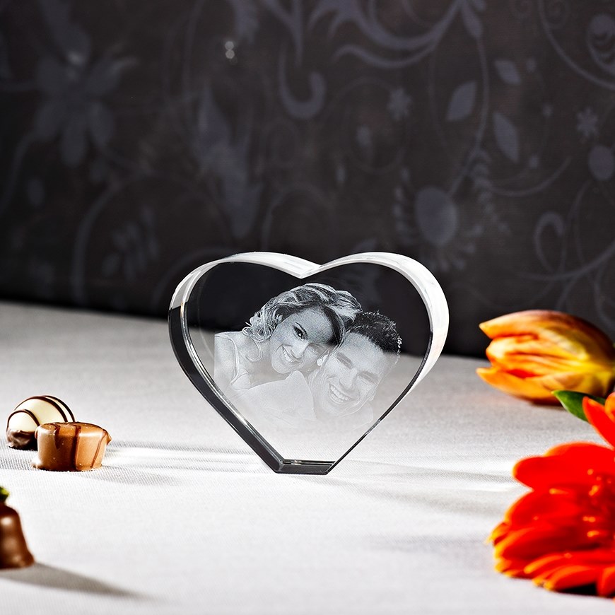 Herzförmiger Kristall mit eingraviertem Paar, umgeben von Pralinen und orangefarbenen Blumen auf weißem Tuch